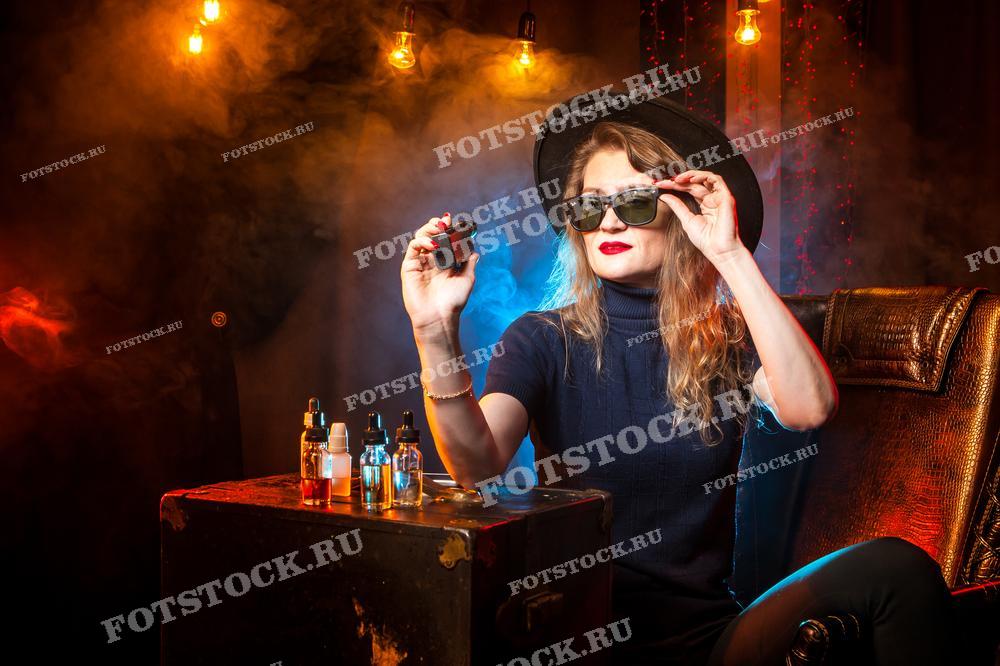 Женщина смотрит на VAPE. На фотографии стильная девушка в солнцезащитных очках демонстрирует электронные сигареты и аксессуары для вейпинга. Женщина курит вейп. Жидкость для электронных сигарет. Vape.
